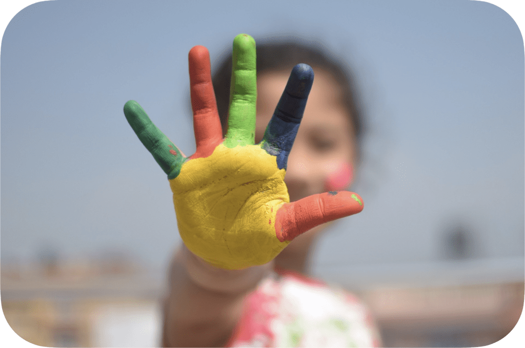 A girl is showing her hand where every finger is painted with a different coloured paint.